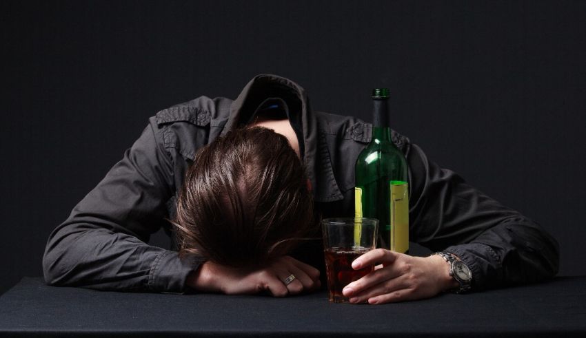 A man with his head down on a table with a bottle of alcohol.