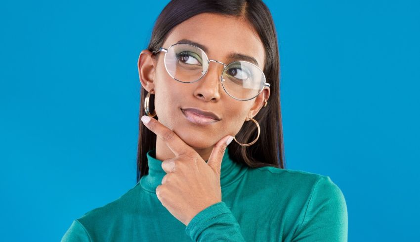 A young woman wearing glasses is posing with her hand on her chin.