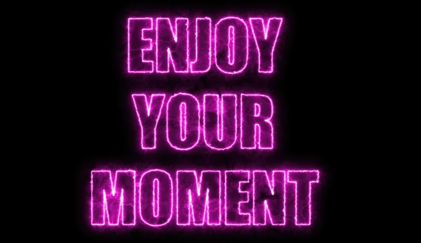 A pink neon sign with the words enjoy your moment on a black background.