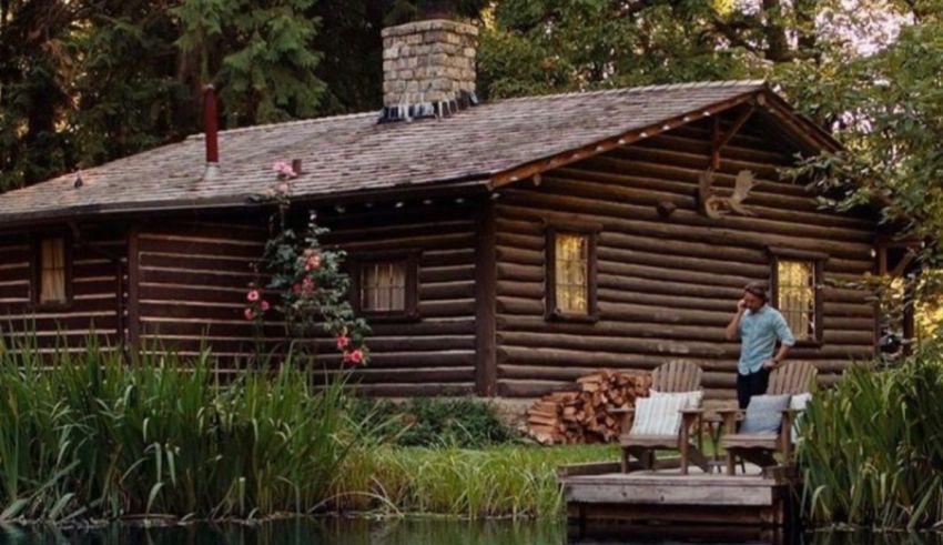 A man sits in front of a log cabin next to a pond.