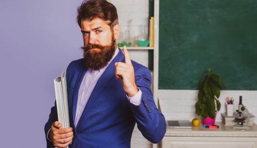 A bearded man with a beard is holding a folder in front of a blackboard.